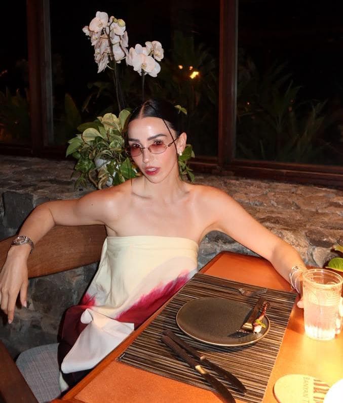 Estíbaliz Badiola posando en un restaurante elegante, con un vestido sin mangas y gafas de sol, rodeada de plantas y flores.