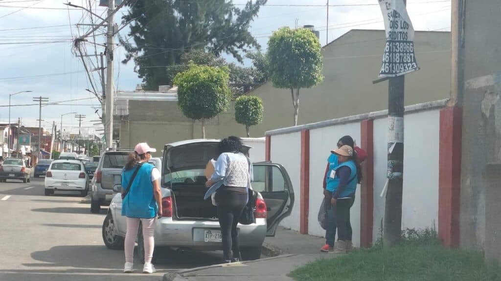 Encuestadores de Morena en Tlaxcala realizando encuestas en la calle, con un vehículo estacionado y personas conversando.